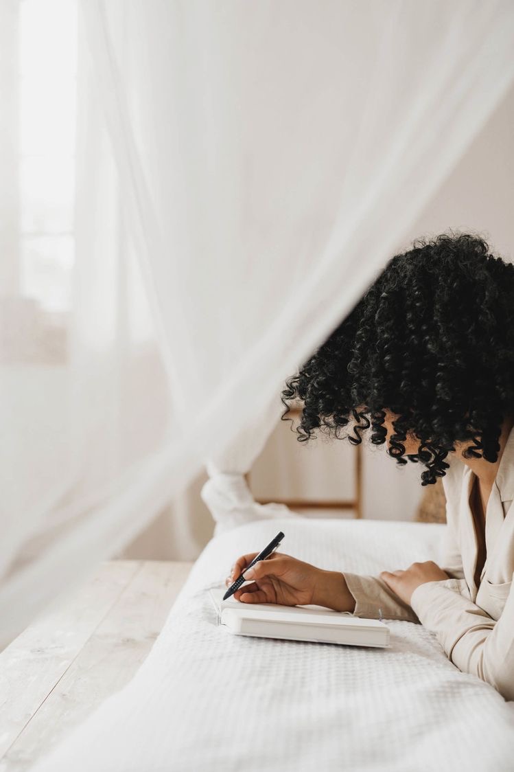 healthy living tips. keep and write in a journal. curly haired woman writing in a journal on the bed