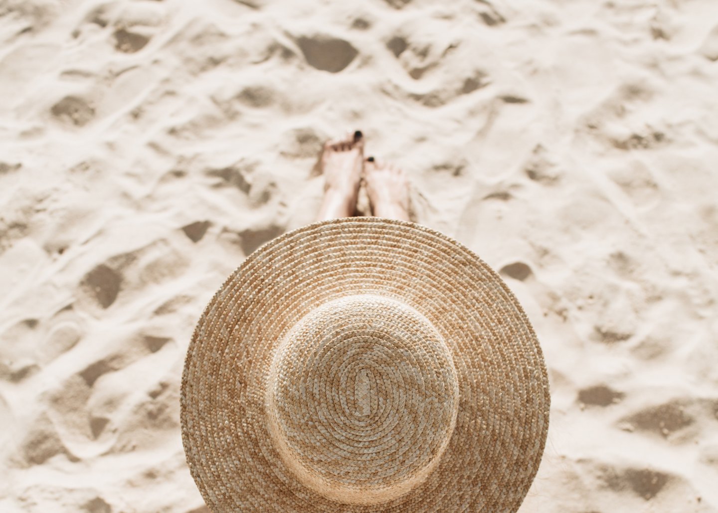 woman with summer hat sitting on the sand on the beach. summer self-care essentials for health and wellness. summer essentials for wellness. best summer wellness items and products.