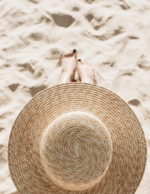 woman with summer hat sitting on the sand on the beach. summer self-care essentials for health and wellness. summer essentials for wellness. best summer wellness items and products.